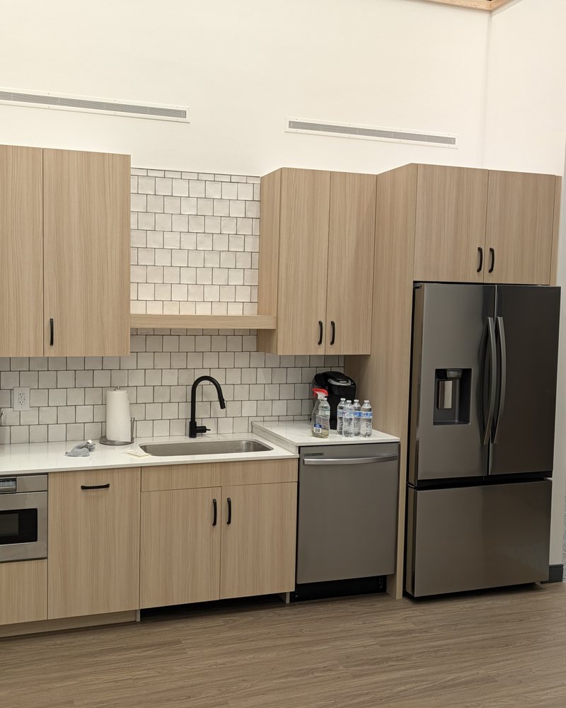 Office break room in light wood-grain laminate with matte black pulls: sink wall, open shelf, tall cabinet around the refrigerator