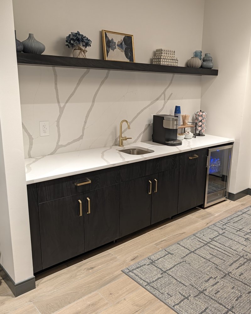 Coffee bar: dark wood-grain laminate slab-front base cabinets with champagne bronze pulls and an undercounter fridge